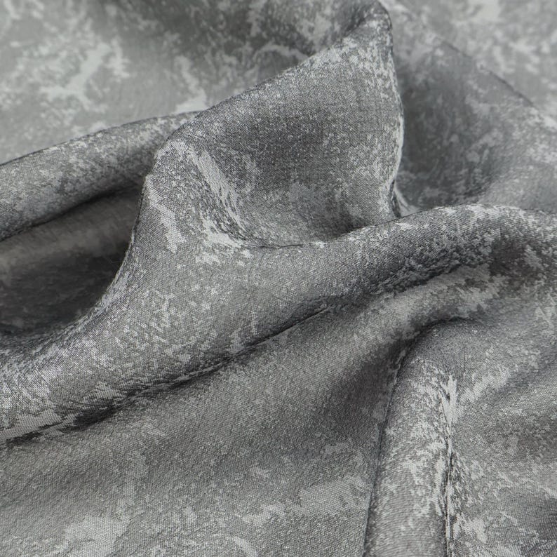 May include: Close-up of a gray fabric with a textured surface and a subtle, abstract pattern in lighter shades of gray and white. The fabric is draped, showing folds and shadows, creating depth and visual interest.