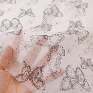 Op de afbeelding: Transparante witte stof met een herhaald patroon van geschetste vlinders in grijstinten en zilver. De stof heeft een delicate, luchtige kwaliteit, met kleine, verspreide zilveren accenten.
