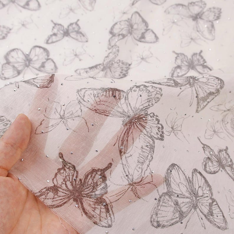 Op de afbeelding: Transparante witte stof met een herhaald patroon van geschetste vlinders in grijstinten en zilver. De stof heeft een delicate, luchtige kwaliteit, met kleine, verspreide zilveren accenten.