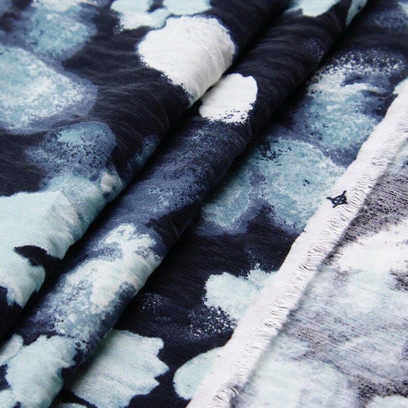 May include: Close-up of a fabric with a watercolor-like pattern. The fabric features a dark navy blue background with large, irregular shapes in white and shades of blue. The fabric is folded, showing the texture and drape.