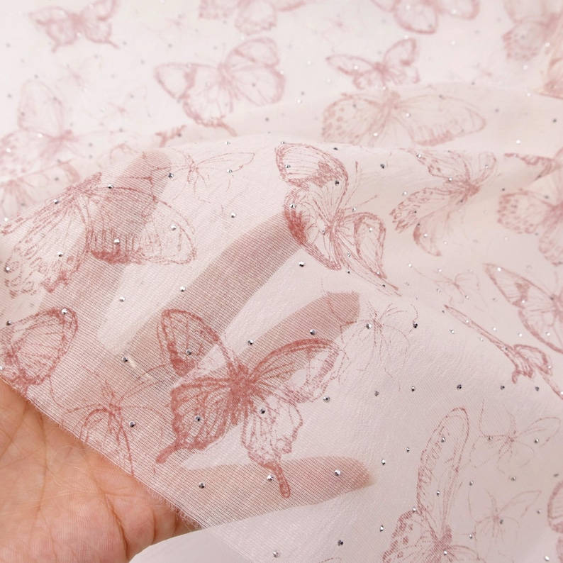 Op de afbeelding: Zachtroze, doorschijnende stof met een patroon van geschetste vlinders. De vlinders zijn in een donkerdere roze tint, met kleine zilveren versieringen verspreid over het oppervlak. De stof is semi-transparant.