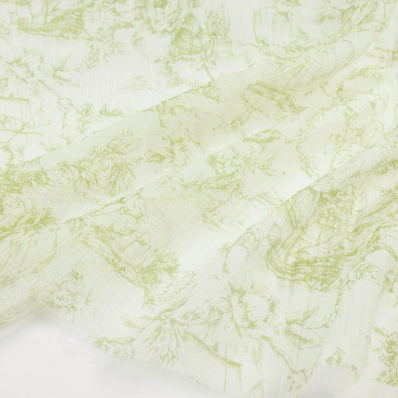 May include: Off-white fabric with a delicate, repeating pattern in light green. The design features a detailed, vintage-style illustration of landscapes, buildings, and floral elements. The fabric has a soft, flowing appearance.
