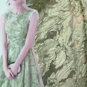 May include: A sleeveless sage green dress featuring a textured, swirling fabric overlay with ruffled details. The garment has a round neckline and a fitted bodice that transitions into a flared skirt, showcasing a layered, dimensional surface pattern.