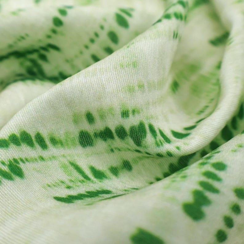 Op de afbeelding: Een zachte, lichtgewicht stof met een witte basis en een levendig groen tie-dye patroon. Het ontwerp bestaat uit horizontale, onderbroken lijnen en verspreide stippen van verschillende groottes, wat zorgt voor een textuurrijk, golvend effect.