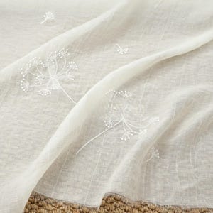 May include: Off-white sheer fabric with embroidered white dandelion designs. The delicate floral pattern is scattered across the flowing material. The fabric has a soft, textured appearance, suitable for curtains or decorative purposes.