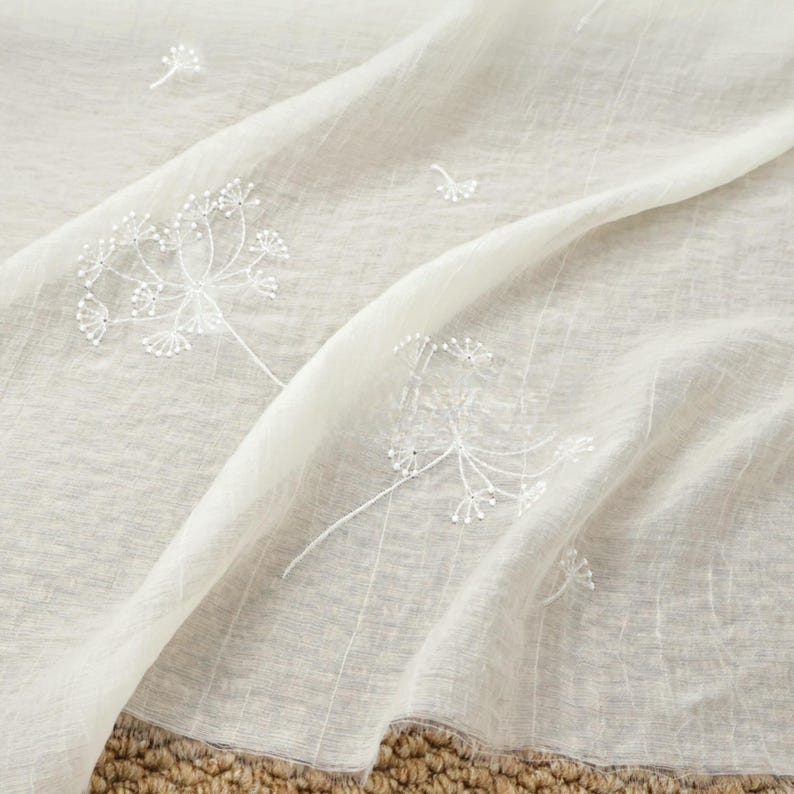May include: Off-white sheer fabric with embroidered white dandelion designs. The delicate floral pattern is scattered across the flowing material. The fabric has a soft, textured appearance, suitable for curtains or decorative purposes.