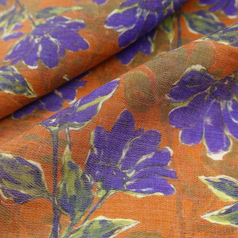 May include: Close-up of a fabric with a floral pattern. The fabric features large purple flowers with green leaves against an orange background. The fabric is folded, showing the texture and depth of the material. Ideal for apparel or home decor.