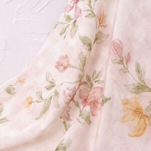 May include: A close-up of a fabric with a watercolor floral pattern. The design features pink, peach, and yellow flowers with green leaves on a pale pink background. The fabric is draped, showing folds and texture.