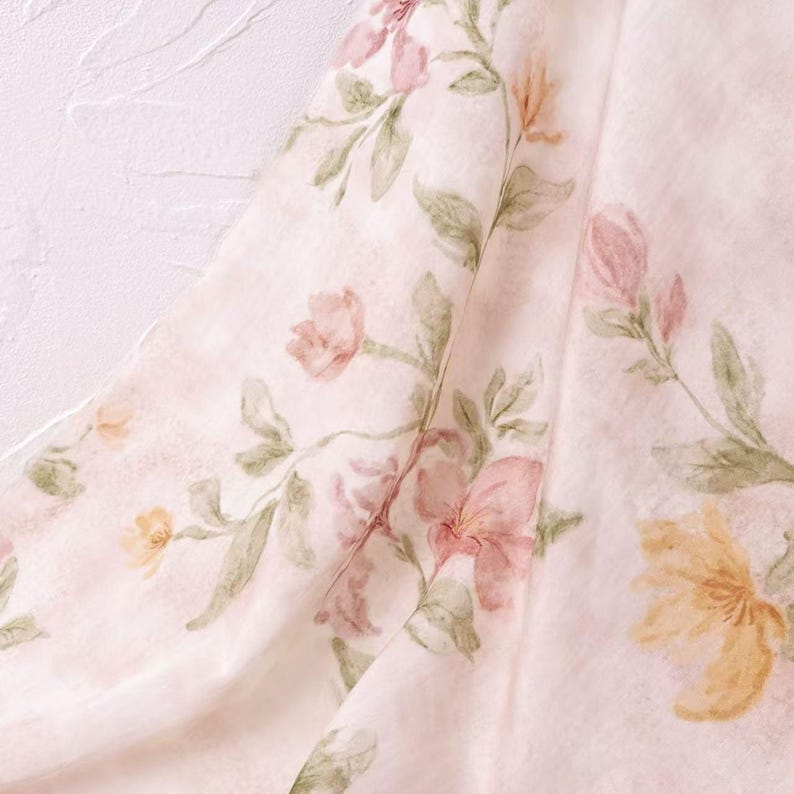 May include: A close-up of a fabric with a watercolor floral pattern. The design features pink, peach, and yellow flowers with green leaves on a pale pink background. The fabric is draped, showing folds and texture.