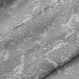 May include: Close-up of a textured fabric with a pattern of light gray and white brushstrokes. The fabric is folded, revealing the texture and the interplay of light and shadow. The overall effect is a soft, artistic design.