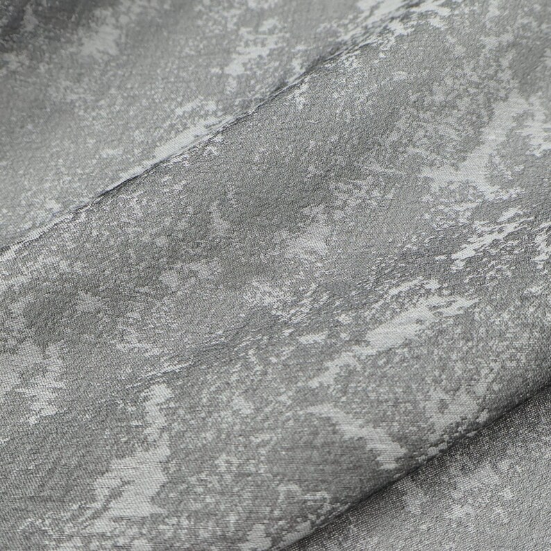 May include: Close-up of a textured fabric with a pattern of light gray and white brushstrokes. The fabric is folded, revealing the texture and the interplay of light and shadow. The overall effect is a soft, artistic design.