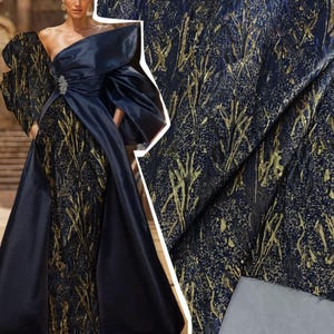 May include: A floor-length gown featuring a dark navy blue satin bodice with a one-shoulder neckline and a silver-toned brooch. The skirt and sleeves are made of a textured fabric with a dark navy blue base and a metallic gold branch-like pattern.