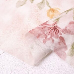 May include: Close-up of a delicate, sheer fabric with a floral pattern. The fabric features a soft pink background with watercolor-style flowers in shades of pink, yellow, and green. The fabric is folded, revealing its layered texture.