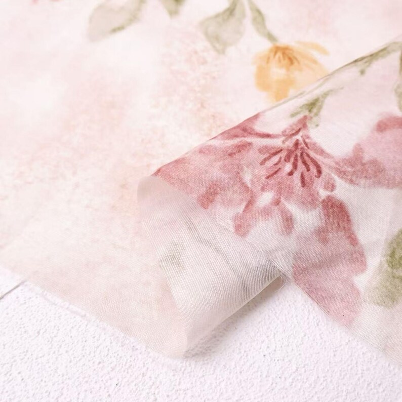 May include: Close-up of a delicate, sheer fabric with a floral pattern. The fabric features a soft pink background with watercolor-style flowers in shades of pink, yellow, and green. The fabric is folded, revealing its layered texture.