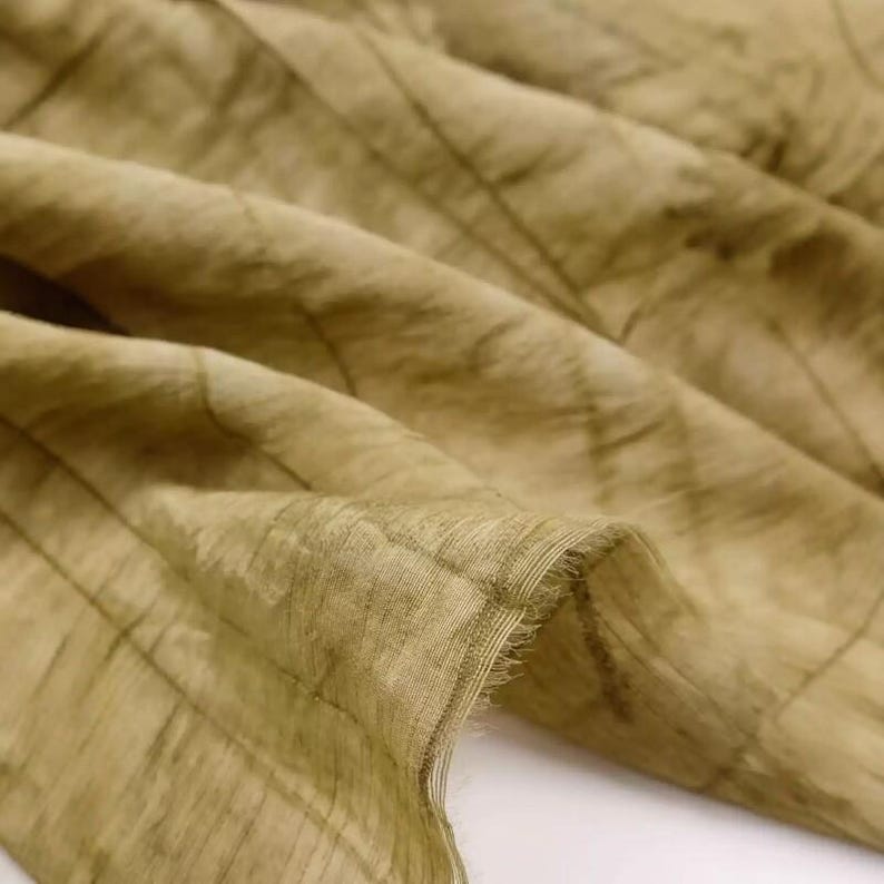 Pode incluir: Uma pe&ccedil;a de tecido semitransparente num tom verde-azeitona suave, com um padr&atilde;o linear subtil e repetitivo. O material apresenta uma textura delicada e enrugada com dobras vis&iacute;veis e uma extremidade crua e desfiada na lateral do tecido.