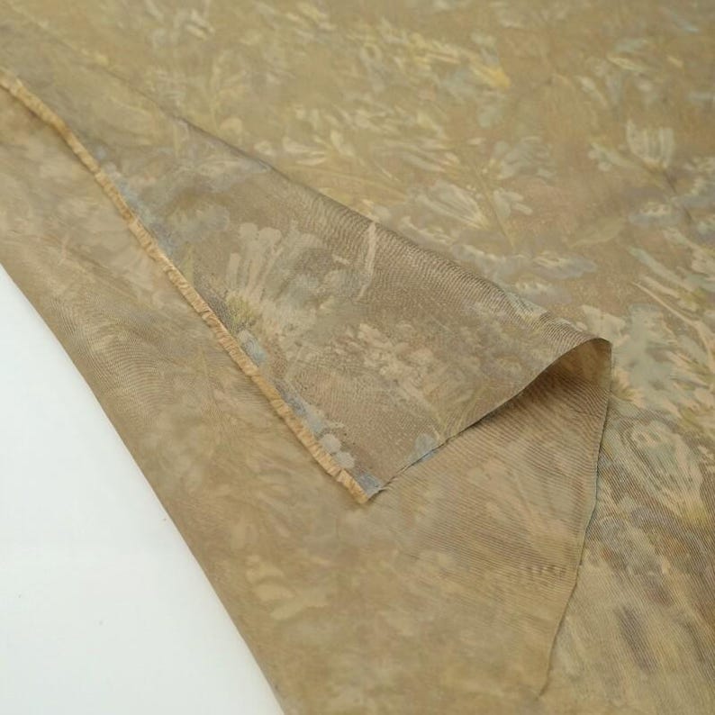 Puede incluir: Una pieza de tela transparente de color marr&oacute;n claro con un sutil patr&oacute;n floral tonal. El material tiene un tejido fino y semitransparente con una textura delicada, mostrando un borde doblado que revela el intrincado dise&ntilde;o bot&aacute;nico del tejido.