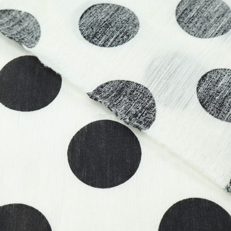 May include: Close-up of white fabric with large black polka dots. The fabric is folded, revealing the texture and pattern. The polka dots vary in texture, some solid and others with a textured, speckled appearance.