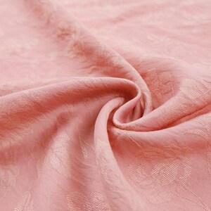 May include: A soft, light pink fabric featuring a subtle, tone-on-tone jacquard weave pattern of roses and leaves. The material is draped in gentle folds, highlighting the textured, embossed floral design across the surface of the textile.