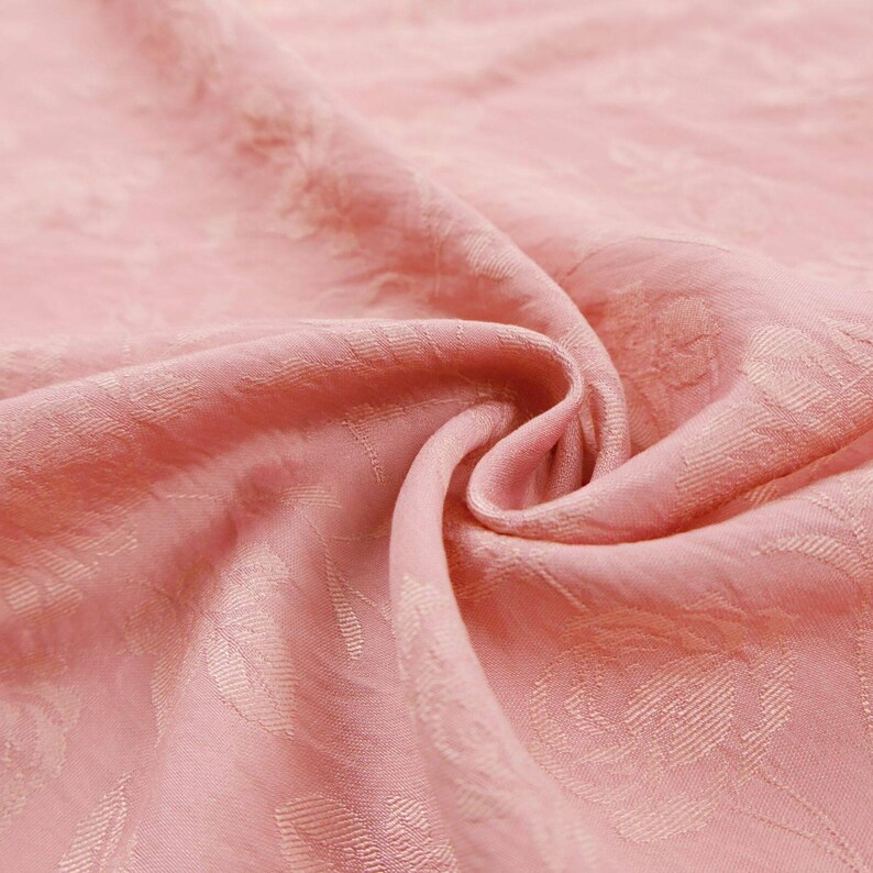 May include: A soft, light pink fabric featuring a subtle, tone-on-tone jacquard weave pattern of roses and leaves. The material is draped in gentle folds, highlighting the textured, embossed floral design across the surface of the textile.