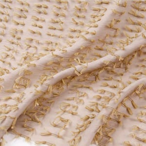 May include: A light beige fabric featuring a repeating pattern of small, raised, fringed tufts in a matching beige tone. The material has a soft, semi-sheer quality with a textured surface created by the evenly spaced, short, frayed thread accents.