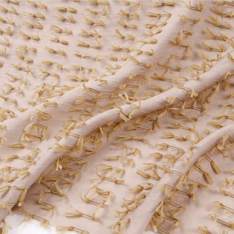May include: A light beige fabric featuring a repeating pattern of small, raised, fringed tufts in a matching beige tone. The material has a soft, semi-sheer quality with a textured surface created by the evenly spaced, short, frayed thread accents.