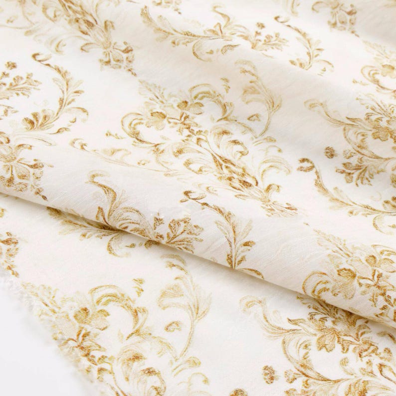 May include: Off-white fabric with a repeating gold floral pattern. The design features stylized flowers and swirling leaves, creating a classic, elegant look. The fabric is folded, showing its texture and drape.
