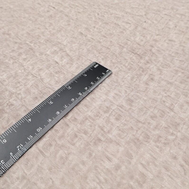 May include: A black ruler with white markings, measuring up to 13 inches, lies diagonally across a textured, light brown surface. The ruler is made of a dark material and features both inch and centimeter measurements.