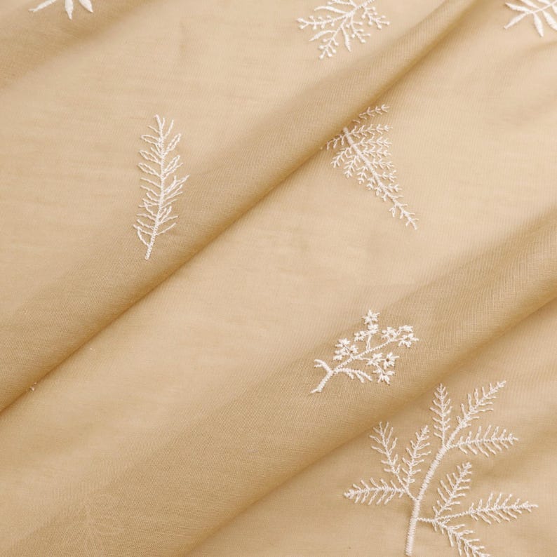 May include: Close-up of beige fabric with white embroidered floral and leaf patterns. The fabric is folded, creating diagonal lines across the image. The embroidery is detailed and delicate, with various botanical designs.