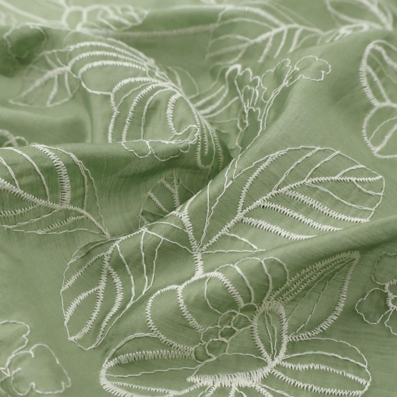 May include: A sage green fabric featuring an intricate white embroidered floral and leaf pattern. The material has a soft, slightly textured surface with visible folds and drapes that highlight the detailed stitching of the botanical design.