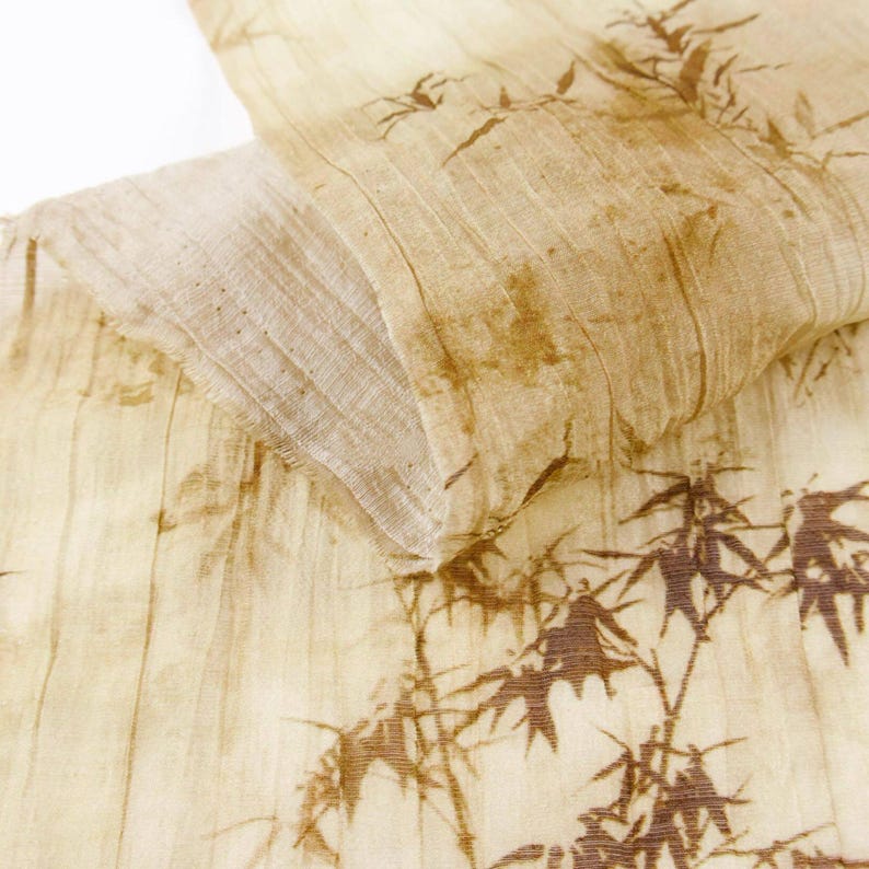 May include: Close-up of a beige fabric with a brown bamboo leaf pattern. The fabric is folded, revealing layers and textures. The design features detailed bamboo stalks and leaves, creating a natural, organic aesthetic. The fabric has a slightly wrinkled appearance.