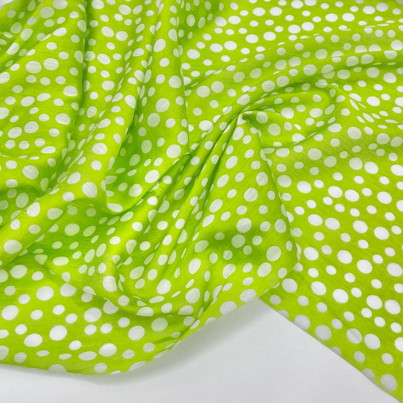 May include: A vibrant lime green fabric featuring a repeating pattern of solid white polka dots of varying sizes. The material is draped in soft folds, creating a textured appearance that highlights the contrast between the bright green and white.
