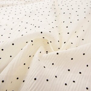 May include: Off-white fabric with a subtle texture and a pattern of evenly spaced black polka dots. The fabric is draped, showing folds and shadows, creating depth. The dots vary slightly in size and are scattered across the surface.