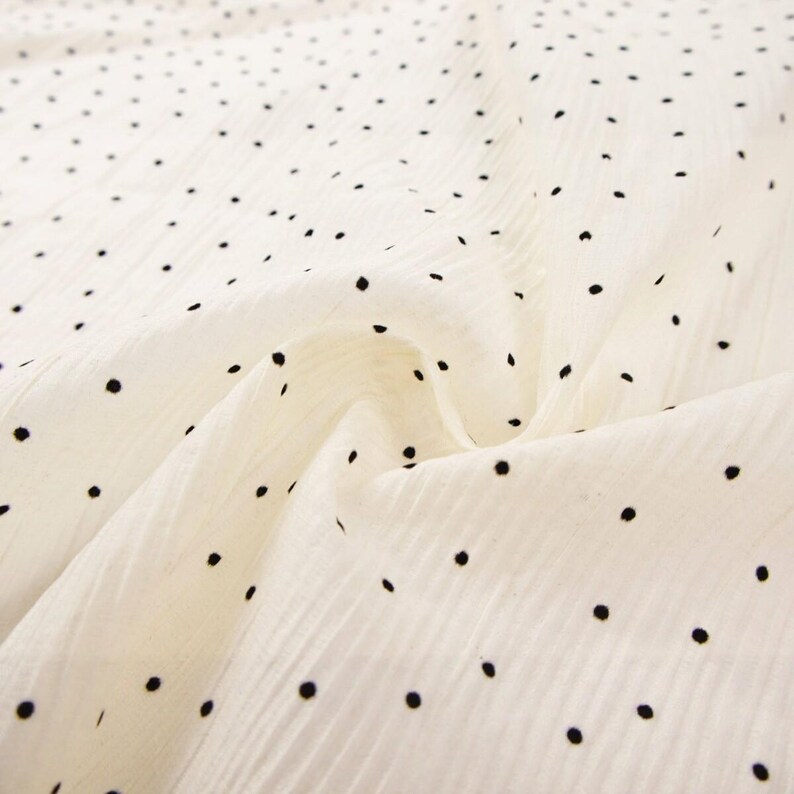 May include: Off-white fabric with a subtle texture and a pattern of evenly spaced black polka dots. The fabric is draped, showing folds and shadows, creating depth. The dots vary slightly in size and are scattered across the surface.