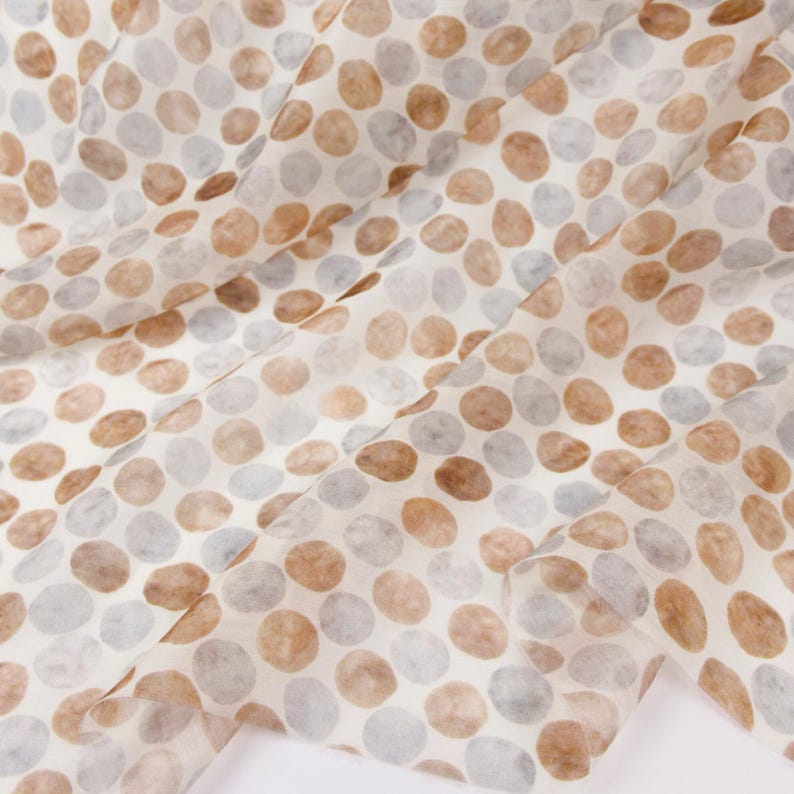 May include: Close-up of a fabric with a repeating pattern of brown and gray circles on a white background. The fabric is folded, creating shadows and depth. The circles vary in size and are evenly spaced.