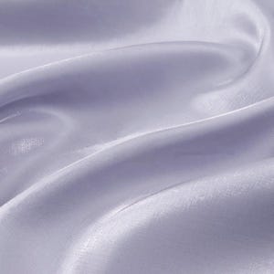 May include: Close-up of a flowing, light lavender fabric. The material has a subtle sheen, with soft folds and gentle curves that create a sense of movement. The fabric appears smooth and delicate, suggesting a lightweight textile.