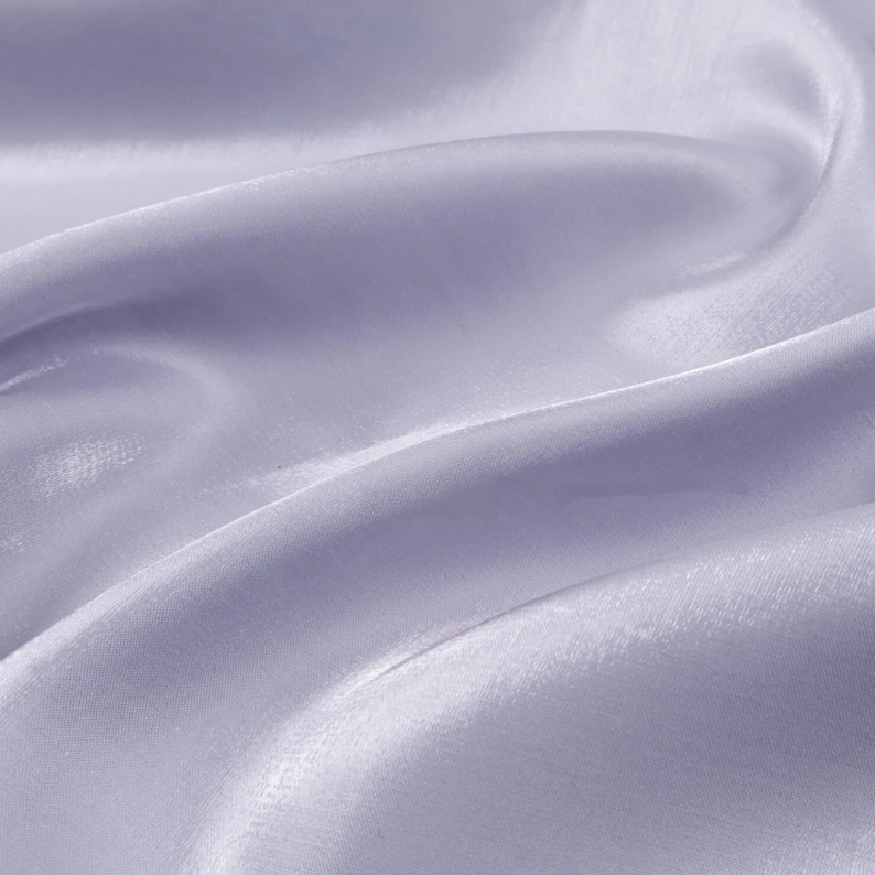 May include: Close-up of a flowing, light lavender fabric. The material has a subtle sheen, with soft folds and gentle curves that create a sense of movement. The fabric appears smooth and delicate, suggesting a lightweight textile.