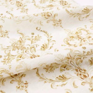 May include: Off-white fabric with a repeating pattern of gold-colored floral and scroll designs. The fabric is folded, showing the texture and detail of the pattern. The design is reminiscent of classic damask.