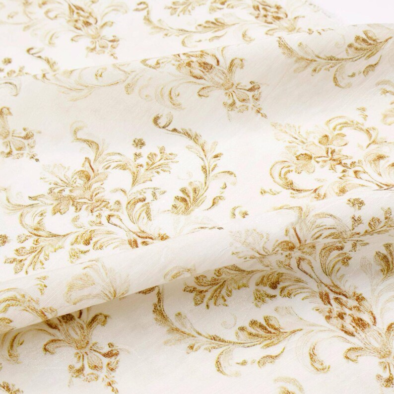 May include: Off-white fabric with a repeating pattern of gold-colored floral and scroll designs. The fabric is folded, showing the texture and detail of the pattern. The design is reminiscent of classic damask.