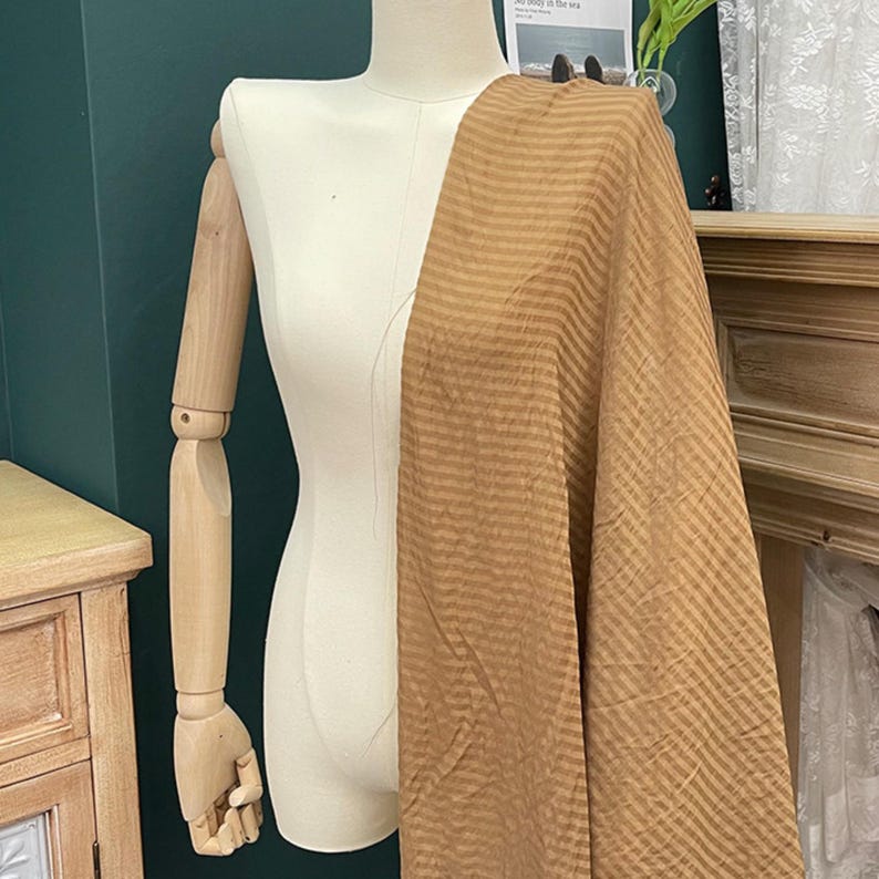 Peut inclure: Une &eacute;charpe beige avec un motif discret de rayures verticales r&eacute;p&eacute;titives drap&eacute;e sur un mannequin couleur cr&egrave;me. Le tissu pr&eacute;sente une texture l&eacute;g&egrave;rement froiss&eacute;e et recouvre le c&ocirc;t&eacute; droit du torse, tombant en plis souples et fluides.