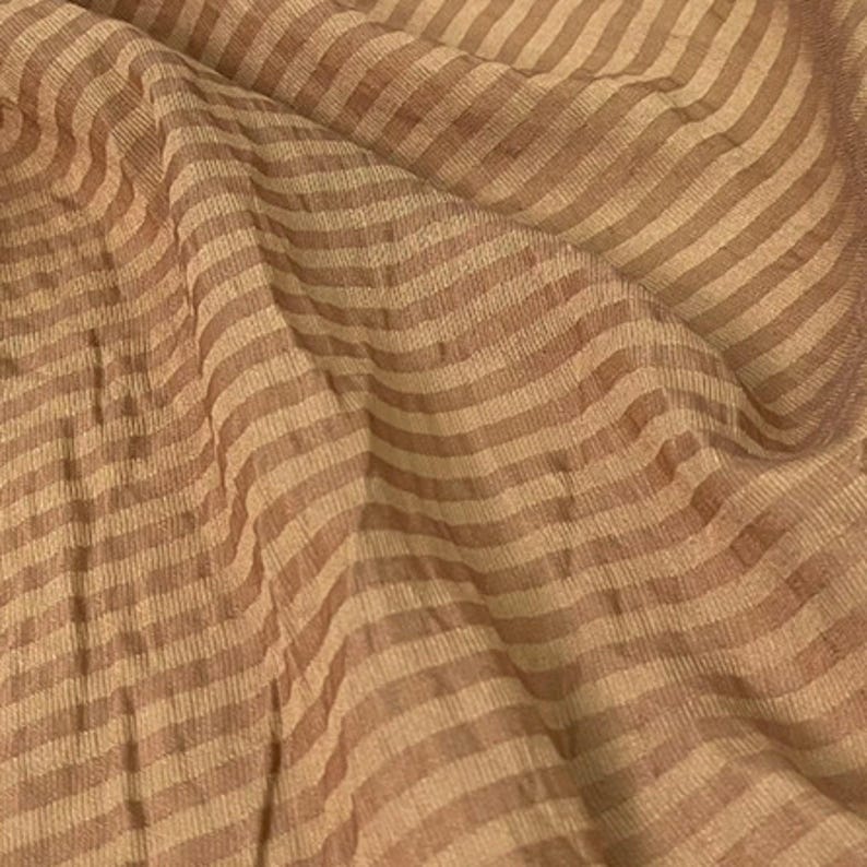 Peut inclure: Un tissu l&eacute;ger pr&eacute;sentant un motif r&eacute;p&eacute;titif de fines rayures horizontales dans des tons altern&eacute;s de beige clair et de brun doux. Le mat&eacute;riau affiche une texture subtilement froiss&eacute;e avec des plis et des drap&eacute;s naturels en surface.