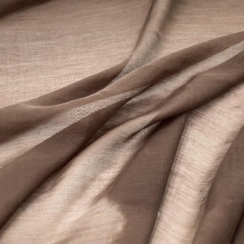 K&ouml;nnte beinhalten: Ein transparenter, leichter Stoff in einem einfarbigen Mokkabraun. Das Material weist ein feines, halbtransparentes Gewebe mit einer subtilen horizontalen Textur auf und ist in weichen, flie&szlig;enden Falten drapiert, die helle und dunkle Braunt&ouml;ne erzeugen.
