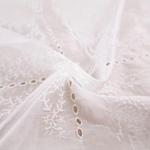 Op de afbeelding: Close-up van witte eyelet stof met ingewikkelde bloemen en geometrische borduursels. Het delicate, vloeiende materiaal is gedrapeerd en toont het gedetailleerde snijwerk en de textuur. Ideaal voor knutselen of kleding.