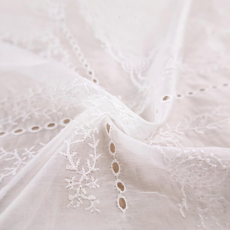 Op de afbeelding: Close-up van witte eyelet stof met ingewikkelde bloemen en geometrische borduursels. Het delicate, vloeiende materiaal is gedrapeerd en toont het gedetailleerde snijwerk en de textuur. Ideaal voor knutselen of kleding.