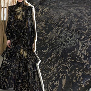 May include: A long-sleeved dress featuring a high neckline and a tiered skirt, crafted from a black fabric with a swirling, marbled pattern in metallic gold and bronze tones. The material displays a textured, liquid-like design throughout the garment.