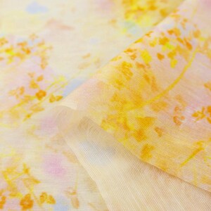May include: Close-up of a sheer fabric with a floral pattern in shades of yellow, orange, and pink. The fabric is folded, revealing a layered texture. The design evokes a watercolor painting, suitable for apparel or home decor.