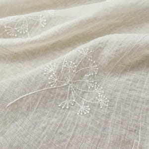 May include: Close-up of a beige fabric with a delicate, embroidered floral pattern. The white embroidery features a detailed, branching design with small, beaded accents. The fabric has a subtle, textured weave, creating a soft, natural aesthetic.