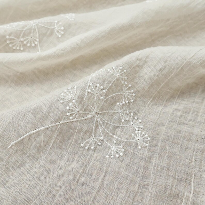 May include: Close-up of a beige fabric with a delicate, embroidered floral pattern. The white embroidery features a detailed, branching design with small, beaded accents. The fabric has a subtle, textured weave, creating a soft, natural aesthetic.