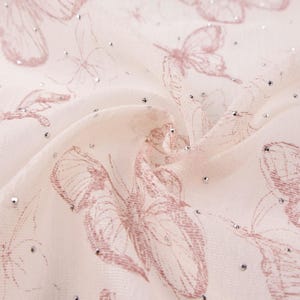 Op de afbeelding: Close-up van een cr&egrave;mekleurige stof met een delicaat vlinderpatroon in een gedempte roze kleur. De stof is versierd met kleine, sprankelende zilveren accenten, wat een glinsterend effect cre&euml;ert. De vlinders zijn ingewikkeld gedetailleerd.