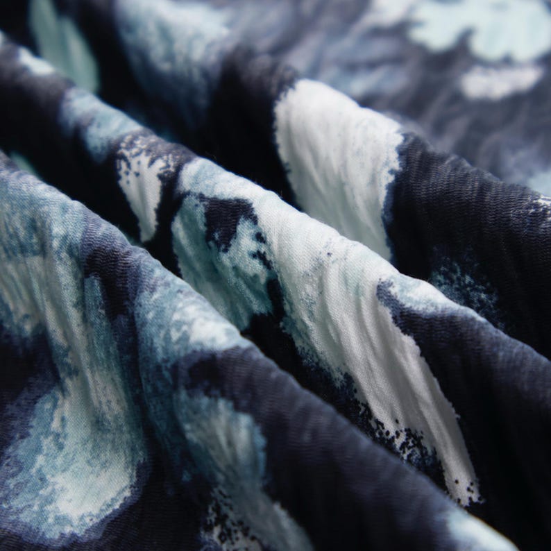 May include: Close-up of a fabric with a watercolor-like pattern. The fabric features a blend of dark navy, light blue, and white hues, creating an abstract design. The material is soft and appears to be draped, with visible folds and textures.
