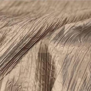 Pode incluir: Close-up de um tecido texturizado com um design plissado. O tecido tem uma aparência brilhante e metálica com uma cor marrom clara. As pregas correm diagonalmente pela moldura, criando uma sensação de profundidade e textura.